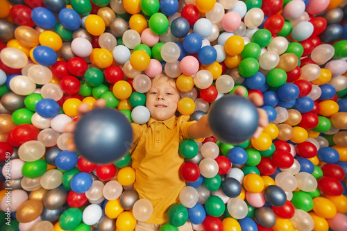 Portrait of happy child lying among colored balls and throwing the balls up