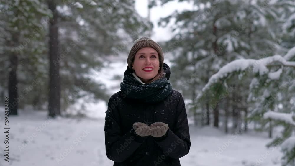Young happy woman in hat and black jacket with red lips snow falls on face smiling in winter park or forest, she catches snowflakes on her gloved hands. Young happy woman in hat and black jacket with red lips snow falls on face smiling in winter park or forest, she catches snowflakes on her gloved hands.