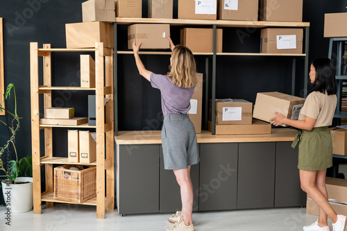 Two young women working in store-room while one of them standing by shelves