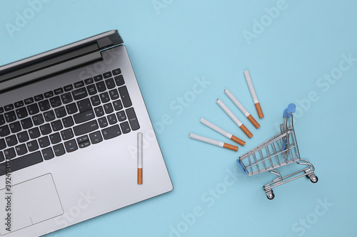 Laptop and shopping cart with cigarettes on a blue background. Top view