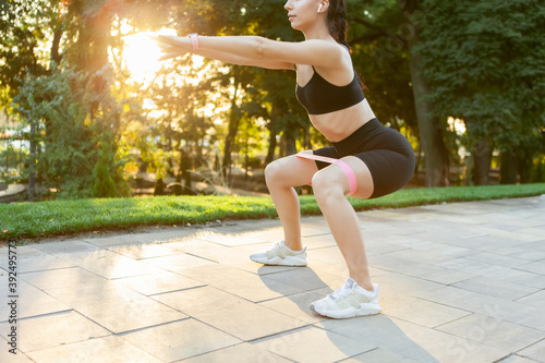 Young attractive brunette woman in sportswear exercises with fitness rubber bands in the park at sunrise