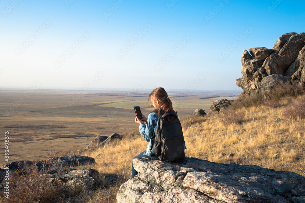A girl with a backpack sits on a rock above the valley in the Golden hour and takes photos. Concept local hiking A girl with a backpack sits on a rock above the valley in the Golden hour and takes photos. Concept local hiking