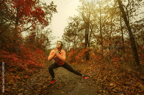Young attractive sport woman in sportswear doing stretching exercise in autumn forest with reddened leaves of trees. Warm up before training. Healthy lifestyle concept