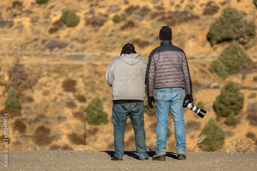 Two men standing on the edge of a road looking into the desert. One holds a large camera