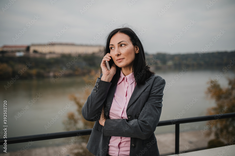 Successful middle age business woman standing on her apartment terrace and talking with someone using her smart phone. Beautiful river view in background. Successful middle age business woman standing on her apartment terrace and talking with someone using her smart phone. Beautiful river view in background.