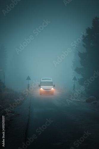 A white van driving on heavy fog with the head lights on a dark moody misty day in the mountains