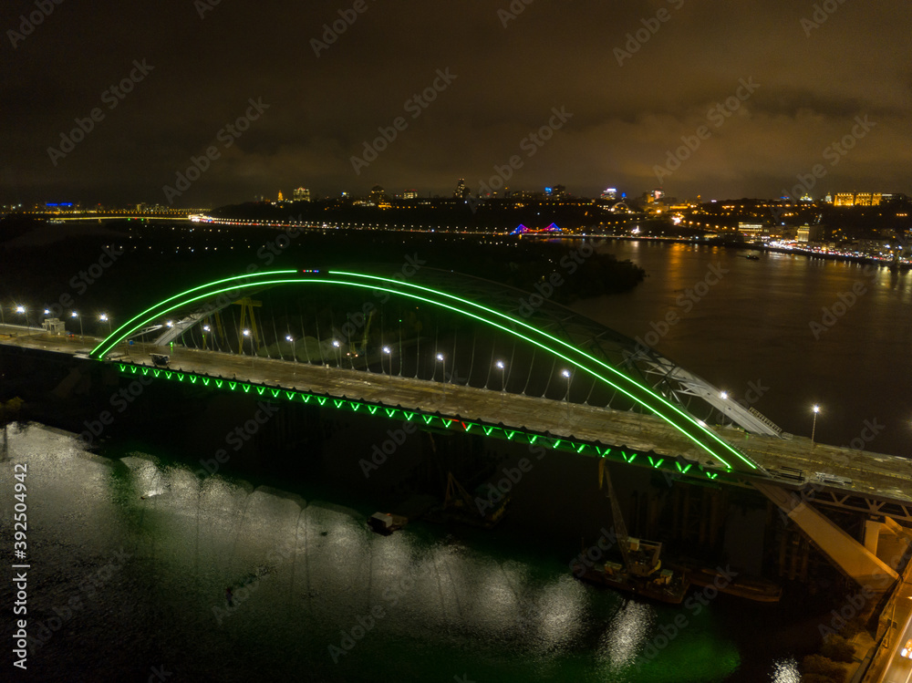 Obraz premium Aerial drone view. Modern arch cable-stayed bridge in Kiev in the evening. Colored lighting of the arch of the bridge.