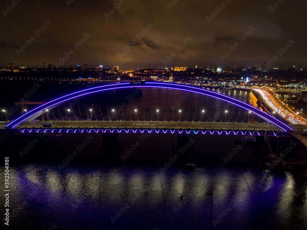 Aerial drone view. Modern arch cable-stayed bridge in Kiev in the evening. Colored lighting of the arch of the bridge. Aerial drone view. Modern arch cable-stayed bridge in Kiev in the evening. Colored lighting of the arch of the bridge.