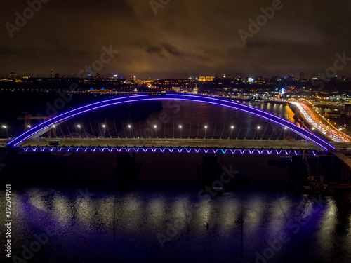 Aerial drone view. Modern arch cable-stayed bridge in Kiev in the evening. Colored lighting of the arch of the bridge. © Sergey