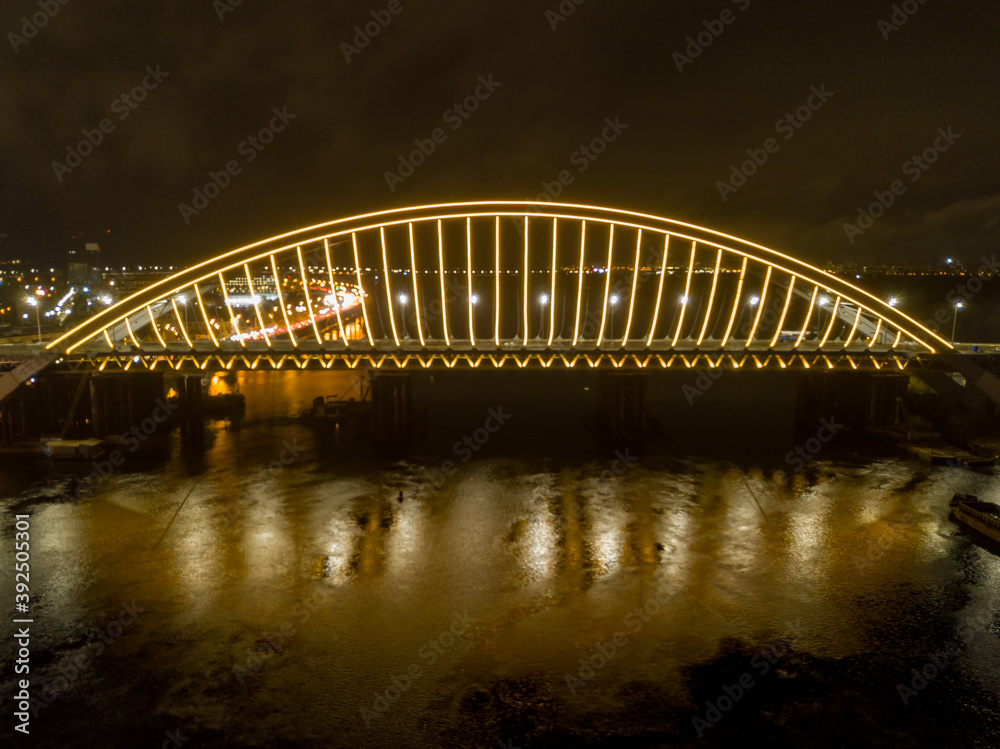 Fototapeta premium Aerial drone view. Modern arch cable-stayed bridge in Kiev in the evening. Colored lighting of the arch of the bridge.