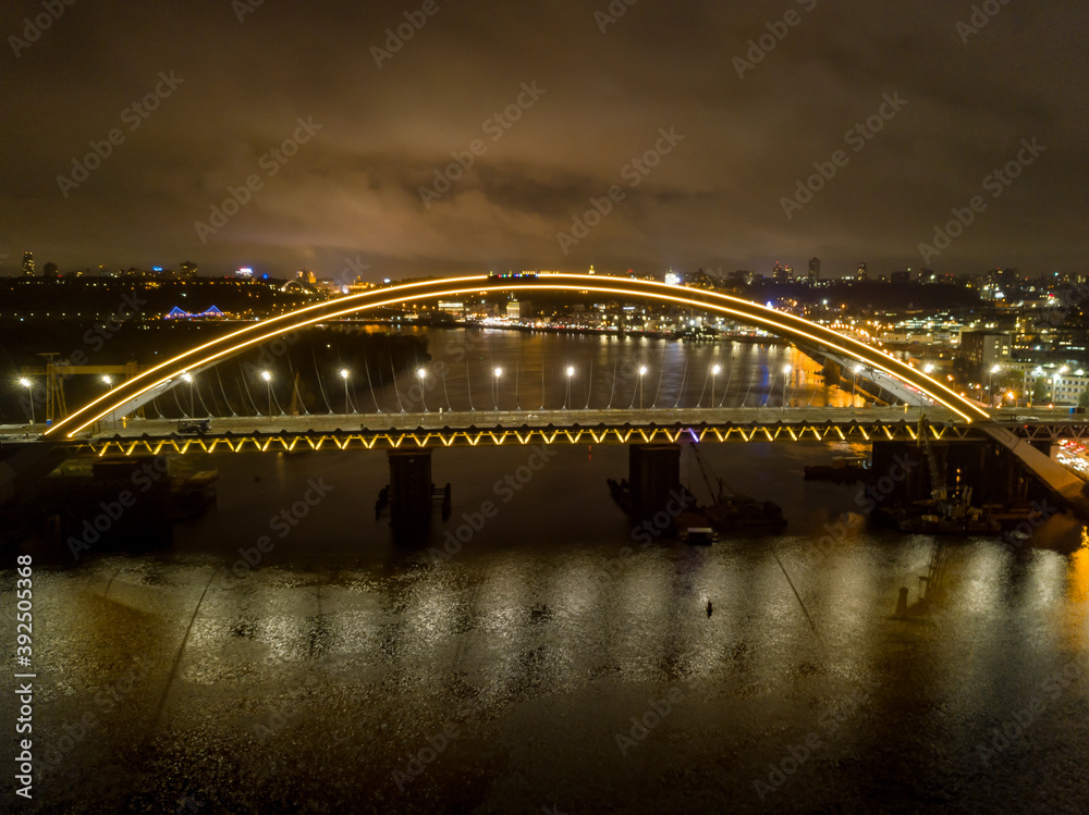 Aerial drone view. Modern arch cable-stayed bridge in Kiev in the evening. Colored lighting of the arch of the bridge.