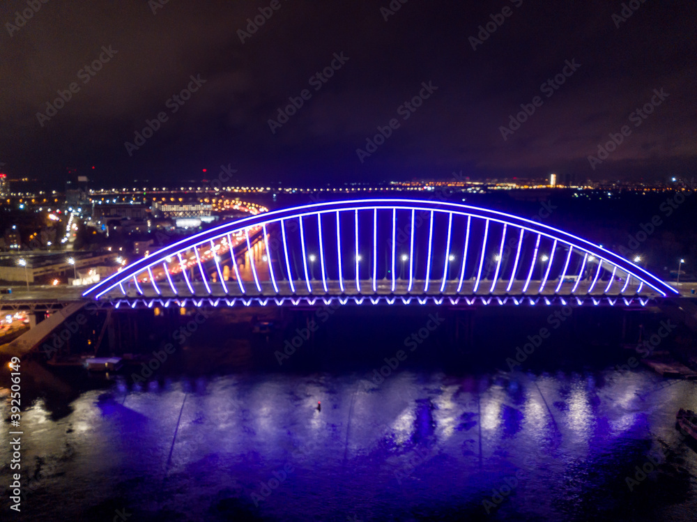 Fototapeta premium Aerial drone view. Modern arch cable-stayed bridge in Kiev in the evening. Colored lighting of the arch of the bridge.
