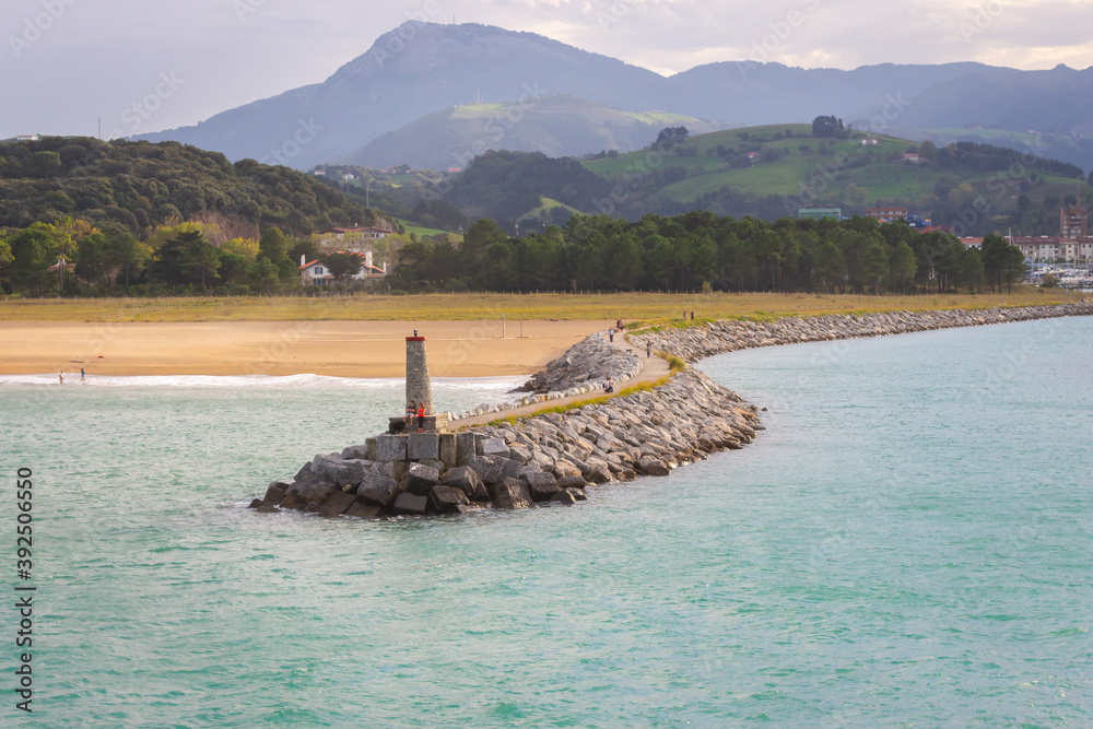 Coast with lighthouse and moor with town and mountains on background. Biscayne bay with scenic coastline. Basque country landscape. Breakwater stones and lighthouse. Commercial dock.