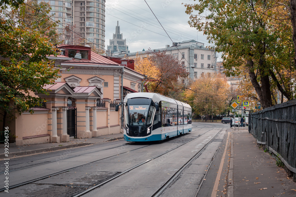 Fototapeta premium A modern tram runs along Dostoevsky Lane. Historical center of Moscow. On the right is an old wooden fence. On the left is an old building. In the background are modern buildings. Fall.
