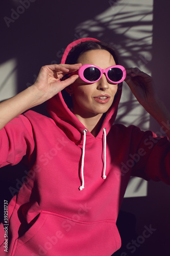 fashionable woman in a pink hoodie and pink sunglasses stands against a wall with a palm tree shadow background in summer
