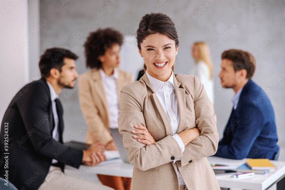 Smiling young businesswoman standing in boardroom with arms crossed and looking at camera. Colleagues in background sitting and working.