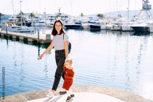 Woman is walking hand in hand with her toddler on a sunny day at a boat marina