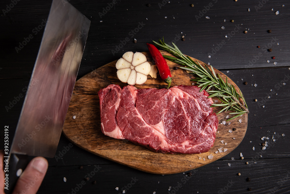 chef holding a knife over fresh juicy marbled beef steak and steak ingredients on cutting board on black wooden table. Chili peppers rosemary garlic top view