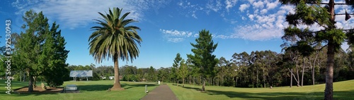 Fototapeta Naklejka Na Ścianę i Meble -  Beautiful panoramic view of a park with green grass and tall trees and deep blue sky with light clouds in the background, Heritage park, Castle Hill, Sydney, New South Wales, Australia
