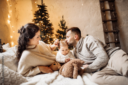 Happy parents with little daughter lay at the bed near decorated Christmas tree, smiling, caring dad gently kiss cute toddler, new year celebration, cozy winter holidays concept