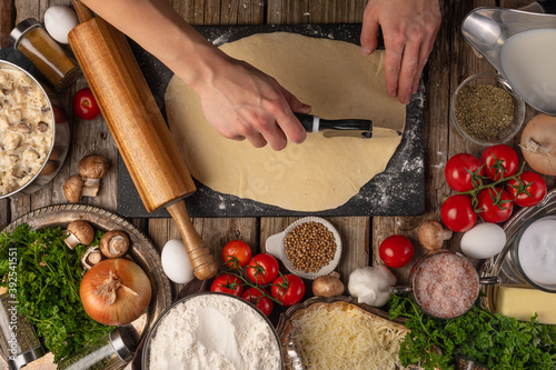 Chef hands cuts with knife the dough for pie on wooden table with variety of ingredients background. Concept of cooking process. Backstage of preparing tasty meal. View from above.