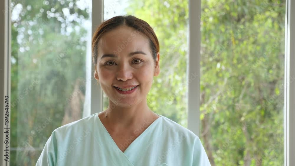 Close up, straight portrait of middle-aged braces Asian female in green patient outfit stand near windows, open eyes, and smile happily among blurred green background of trees. Close up, straight portrait of middle-aged braces Asian female in green patient outfit stand near windows, open eyes, and smile happily among blurred green background of trees.