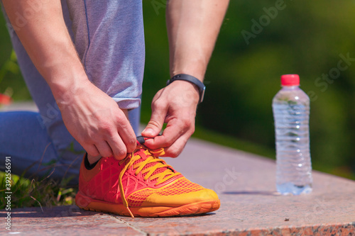 Close up on mans hands tying shoelaces on orange sneakers and a bottle of water during a run in a summer park
