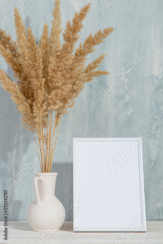 A vase with yellow spikelets and a white photo frame on the table. Light stylish background. Preparation for inscriptions, copy space.