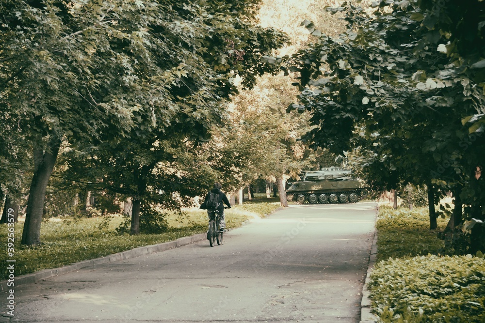 A shady alley in the park with a cyclist on it and overlooking the Soviet tank monument at the end of it. Rybinsk, Russia. August 22, 2020.