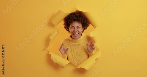 Shocked African American woman suddenly appears in torn paper hole of yellow background raises palms and tries to frighten someone looks gladfully dressed casually. Female tries to be spooky