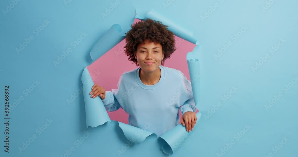 Brunette ethnic woman looks scrupulously at camera tries to see all details nods head with agreement has talk with someone wears casual jumper poses throguh paper blue background. Human reactions