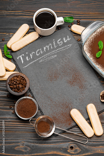 Traditional Italian Tiramisu dessert in glass baking dish, cup of coffee and savoiardi cookies on stone serving board with chalk inscription sign on wooden background