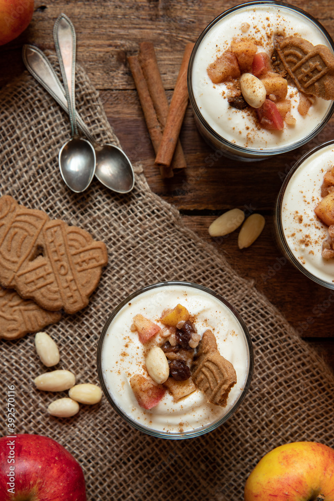 Kuchen im Glas mit Stücken vom Bratapfel, Zimt, Spekulatius, Apfel, Mandeln und Rosinen mit einer Creme aus Quark oder Mascarpone auf einem Tisch aus Holz in einer gemütlichen Küche zu Weihnachten