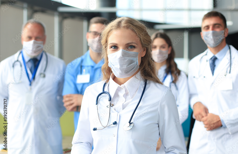 Fototapeta premium Female doctor in medical mask with group of happy successful colleagues also in preventive masks.