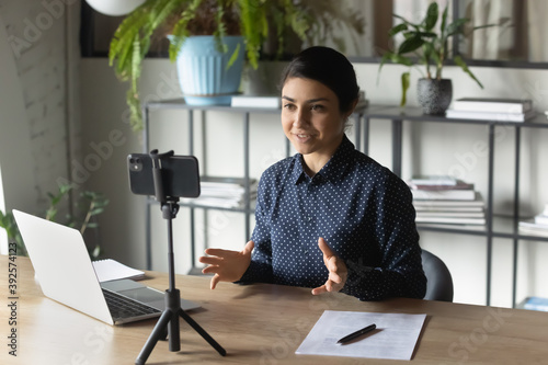 Successful Indian businesswoman recording webinar, using smartphone on tripod, sitting at desk in office, smiling young employee business coach teacher influencer shooting video for blog, speaking photo