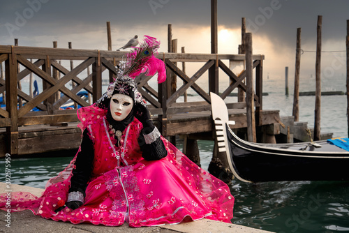 Woman in mask wearing ornate colorful pink carnival costume sits at San Marco lagoon with floating gondolas background at sunset. Carnival in Venice, Italy