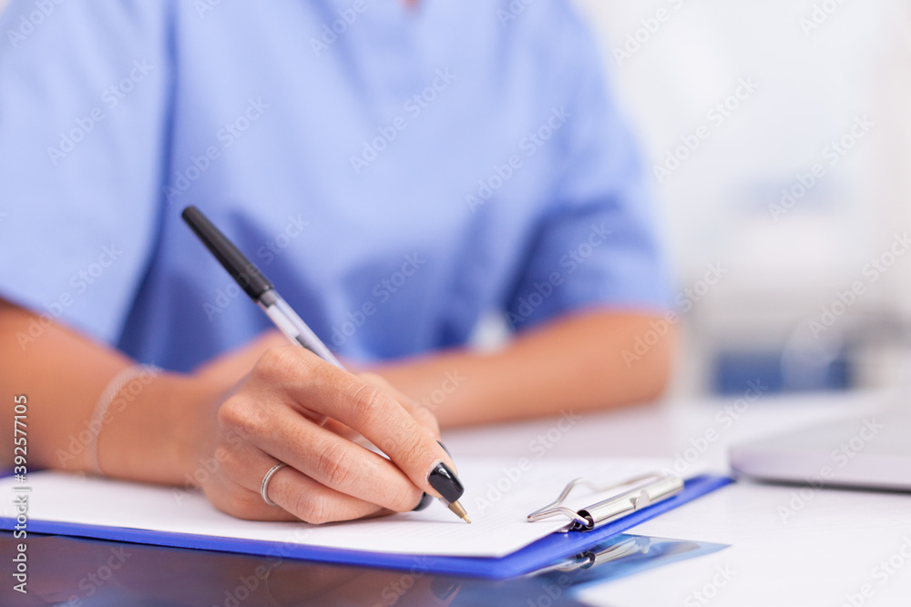 Close up of nurse writing patient diagnosis on clipboard in hospital office . Health care physician using computer in modern clinic looking at monitor, medicine, profession, scrubs.