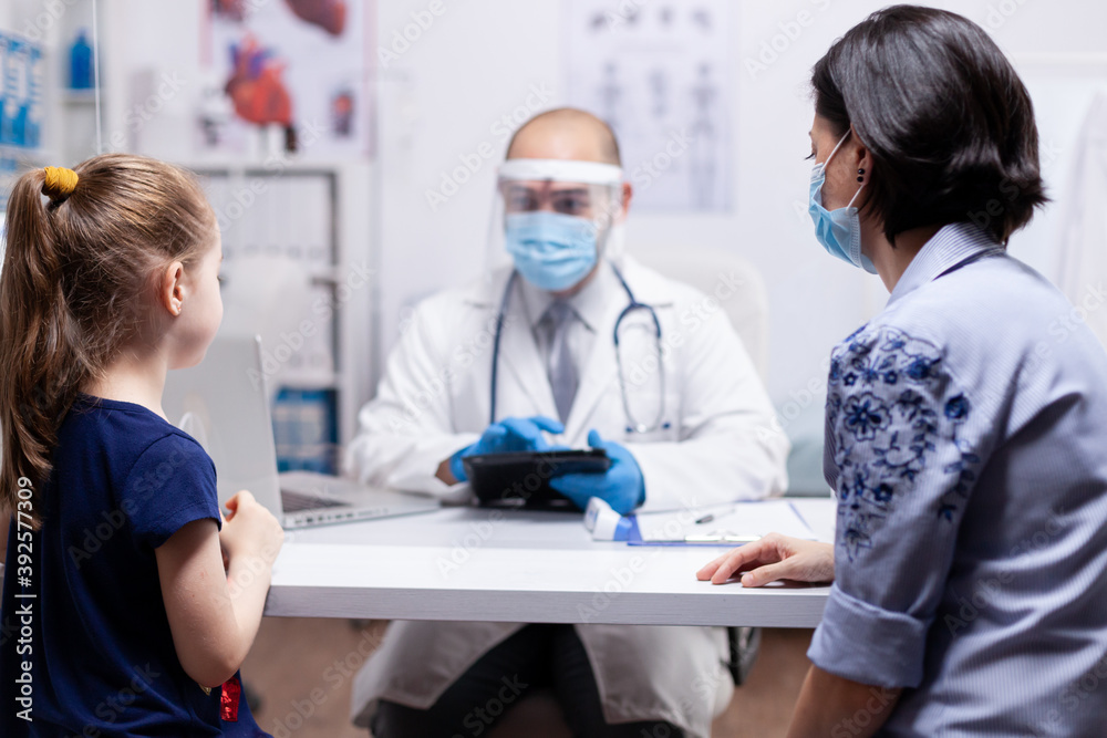 Pediatrician wearing face mask holding tablet pc while consulting child during clobal pandemic with coronavirus. Pediatrician specialist with protection mask providing health care service.