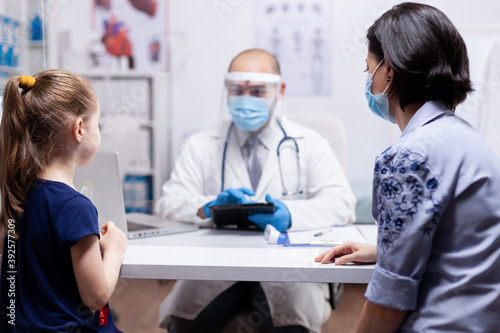 Pediatrician wearing face mask holding tablet pc while consulting child during clobal pandemic with coronavirus. Pediatrician specialist with protection mask providing health care service.