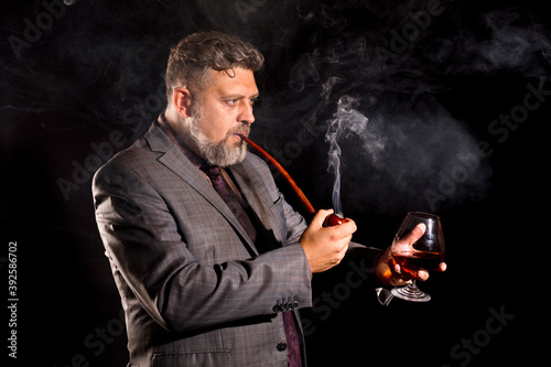 Solid confident bearded man in suit with glass of whisky smoking pipe