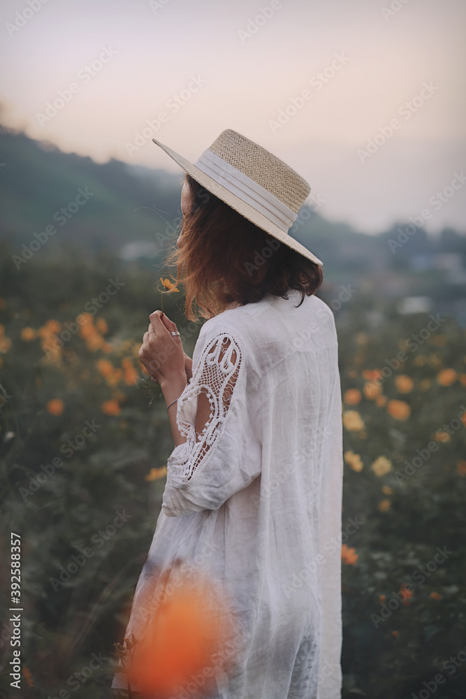 brunette woman in a linen dress and straw hat walks among herbs and wildflowers on a meadow in the mountains. A woman in the Boho style, resting in the countryside, a simple village lifestyle