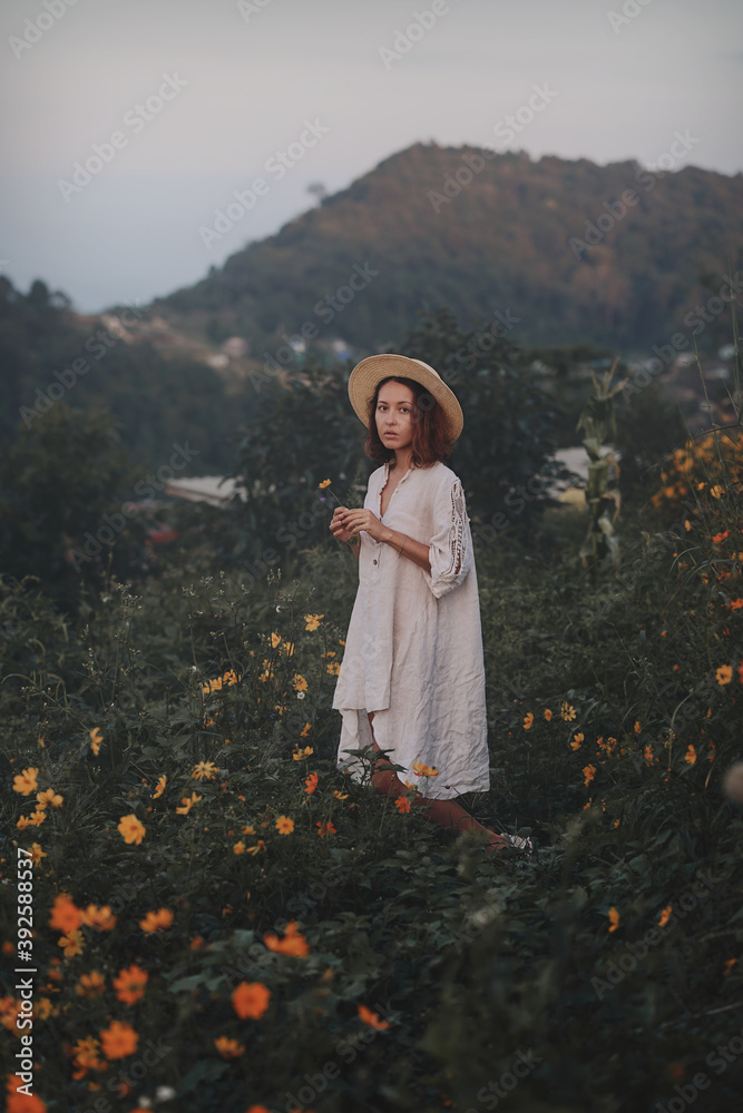 brunette woman in a linen dress and straw hat walks among herbs and wildflowers on a meadow in the mountains. A woman in the Boho style, resting in the countryside, a simple village lifestyle
