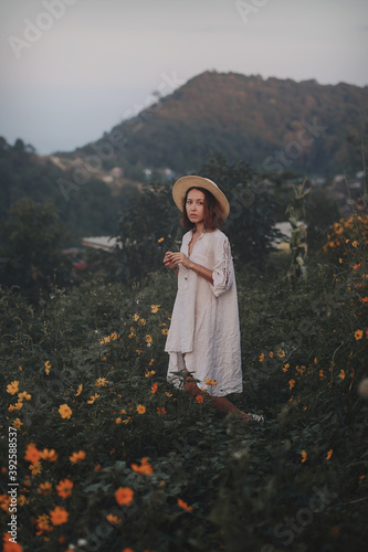 Obraz na płótnie brunette woman in a linen dress and straw hat walks among herbs and wildflowers on a meadow in the mountains. A woman in the Boho style, resting in the countryside, a simple village lifestyle w salonie