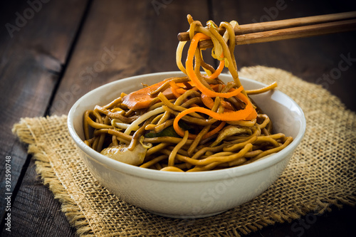 Chopsticks take chinese vegetarian noodles, served in a white bowl on rustic wooden table