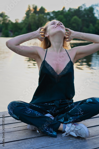 Beautiful blond young woman in dark green lace top pajamas pants sitting near the river bank with forest reflection in the water on blur background 