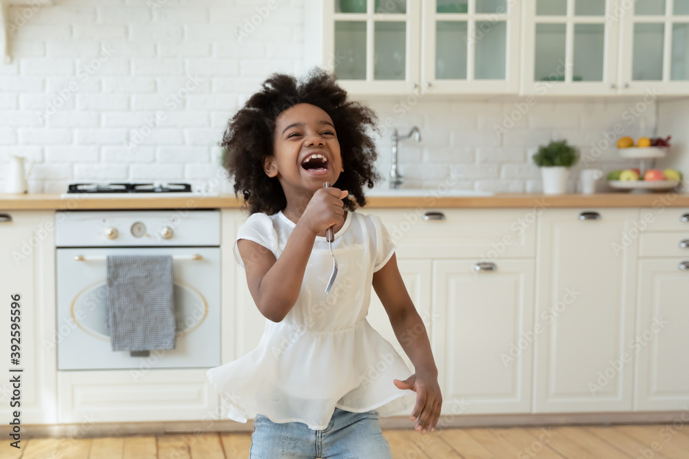 Overjoyed little African American girl holding fork as microphone and singing, standing in modern kitchen, having fun with kitchenware at home, cute child playing funny game, listening to music Overjoyed little African American girl holding fork as microphone and singing, standing in modern kitchen, having fun with kitchenware at home, cute child playing funny game, listening to music
