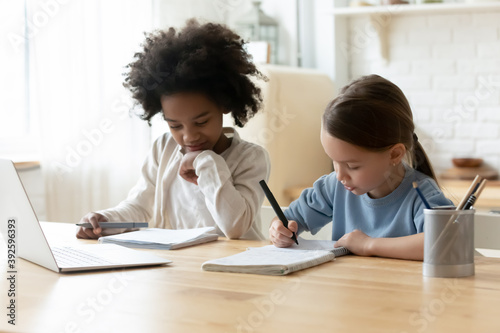 Two diverse pretty little girls studying online at home together, sitting at table in kitchen, multiracial sisters doing school homework, writing notes, listening to lecture, homeschooling concept