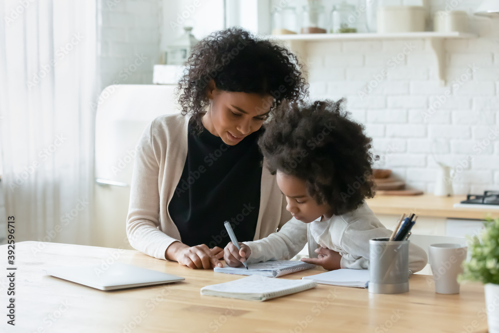 Smiling caring African American mother helping to adorable little daughter with homework, sitting at table in modern kitchen, pretty girl doing school tasks, studying, writing with pen, homeschooling Smiling caring African American mother helping to adorable little daughter with homework, sitting at table in modern kitchen, pretty girl doing school tasks, studying, writing with pen, homeschooling