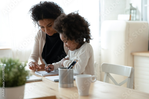 Caring African American mother helping to little daughter with homework, doing school tasks together, sitting at table in modern kitchen, pretty girl writing or drawing, homeschooling concept
