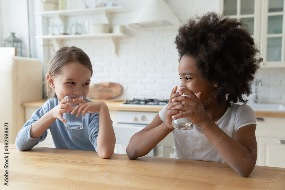 African American and Caucasian cute little girls drinking pure water together, sitting at wooden table in kitchen, pretty kids holding glasses of fresh aqua, refreshment and hydration concept - obrazy, fototapety, plakaty 